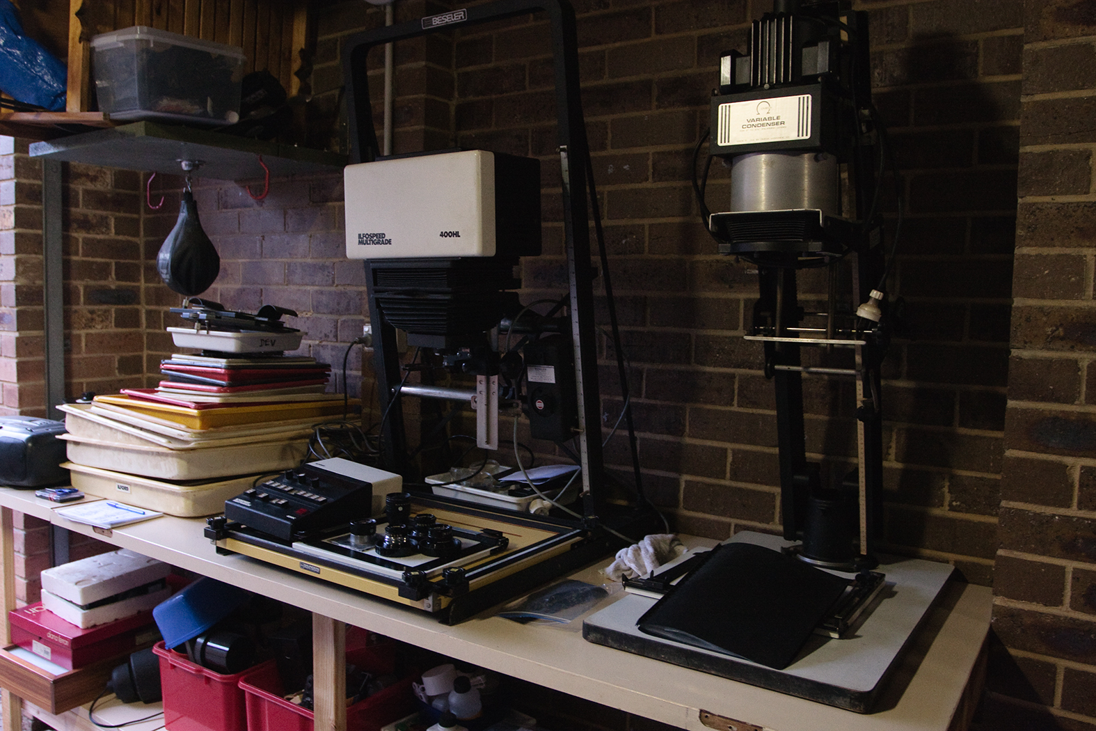 Two photography film enlargers side by side on a worktop desk. There's a collection of trays for processing beside them